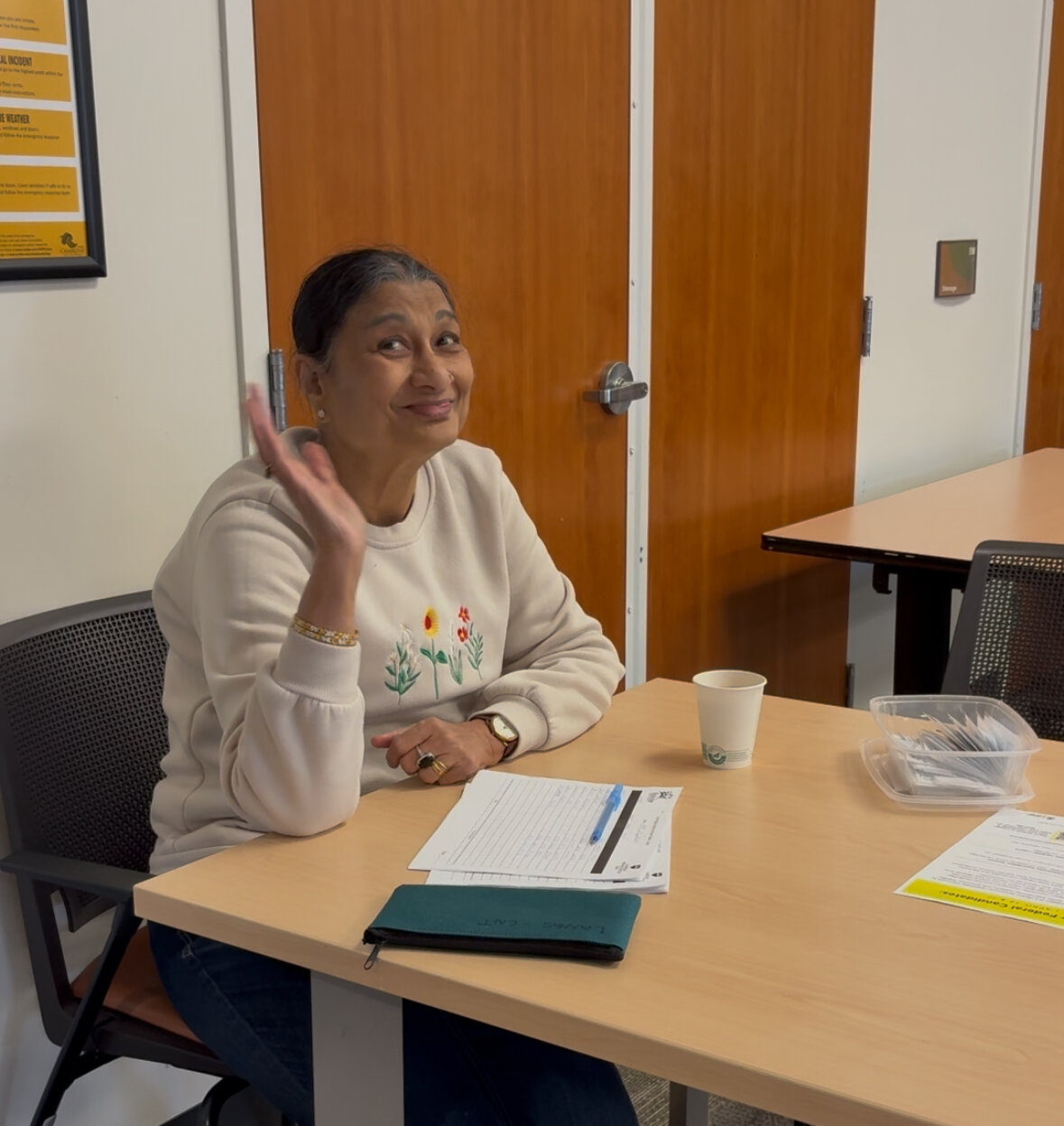 an older brown woman waving while sitting at the sign in table with some papers in front of her